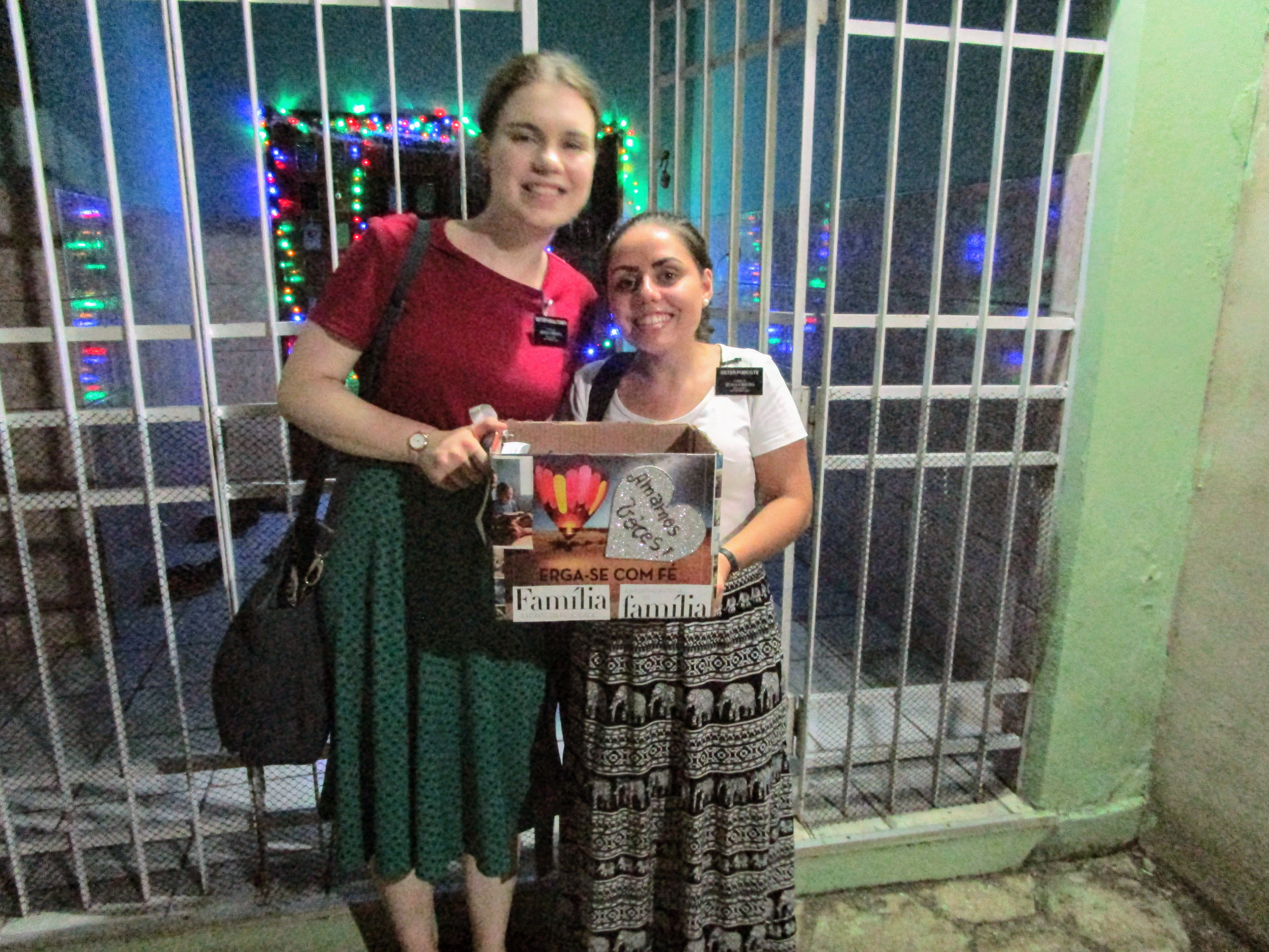 Sister Faulconer and Sister Porcote, Sister missionaries for the Church of Jesus Christ of Latter-day Saints are holding a box of food and other things they put together for a family who needed help. In the background, the green, red and purple Christmas lights they strung up around their apartment are visible. 